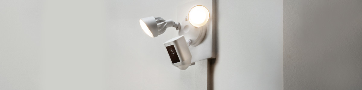 Ring Floodlight Cam (1st Generation) mounted to a white wall outside, with its bright, motion-activated LED lights turned on.